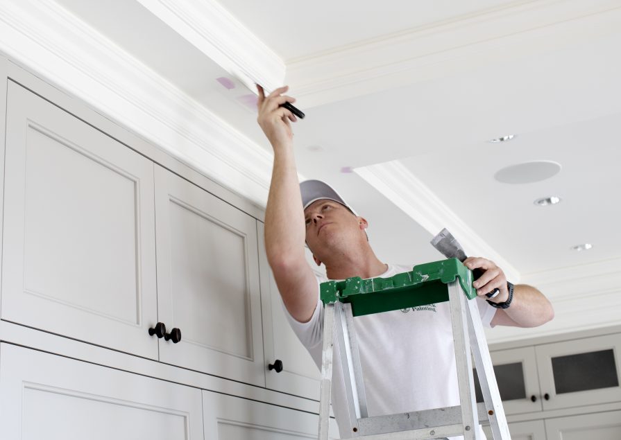 A man dressed in all white stands on a ladder while applying putty to a ceiling