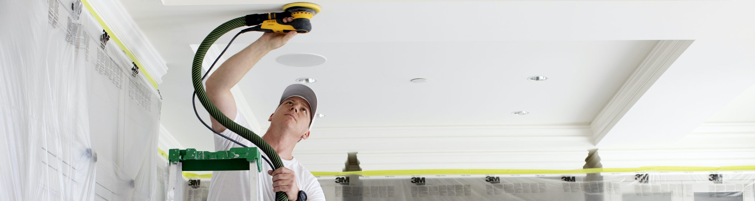 A man standing on a ladder uses a zero-dust sander on a white ceiling