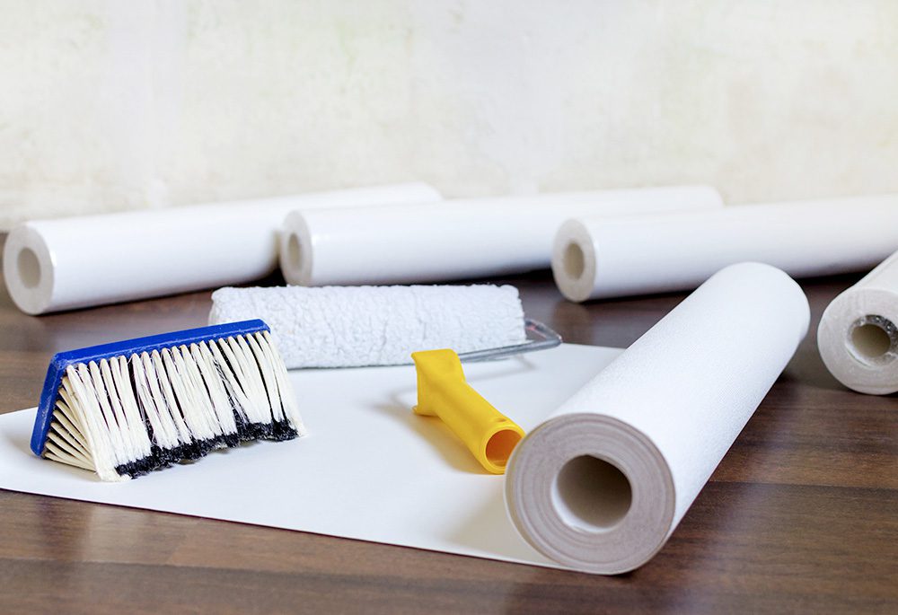 Rolls of wallpaper, a rolling paintbrush, and a wallpaper brush lie on a wooden floor