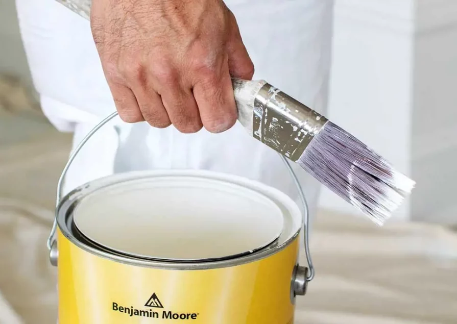 A hand holding a paintbrush and can of white paint