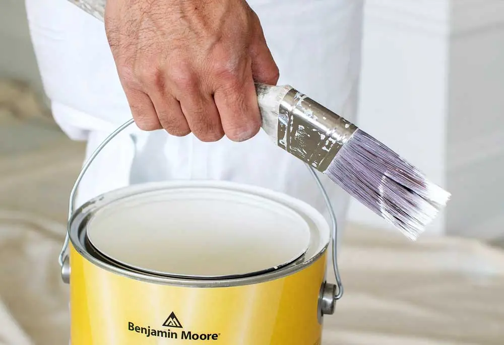 A hand holding a paintbrush and can of white paint